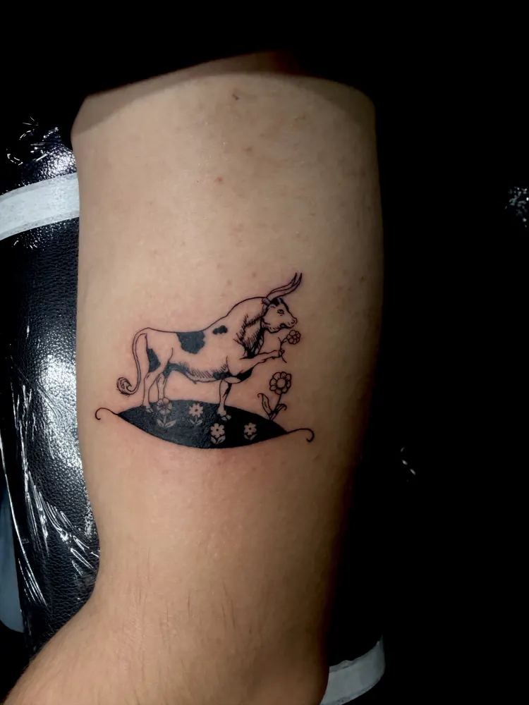 Spotted cow tattoo with a flower and daisies in black linework on the upper arm