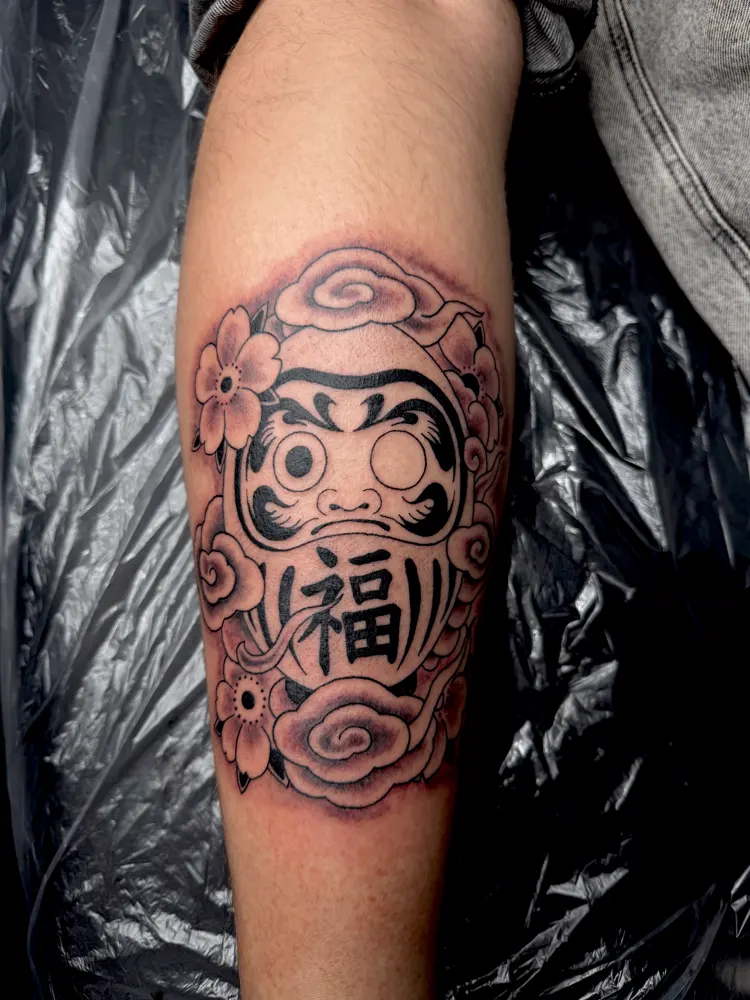 Daruma tattoo with the kanji for good fortune framed by pink flowers and clouds on the shin