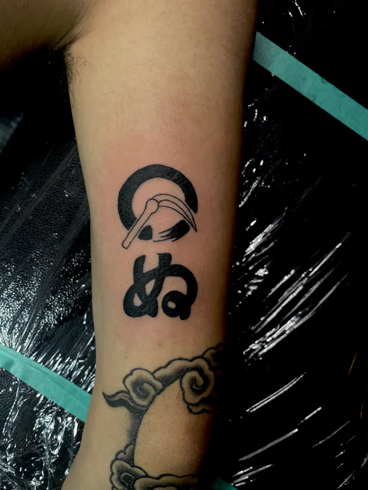 Kamawanu rebus tattoo of a sickle, a circle and the kana nu in solid black on the forearm