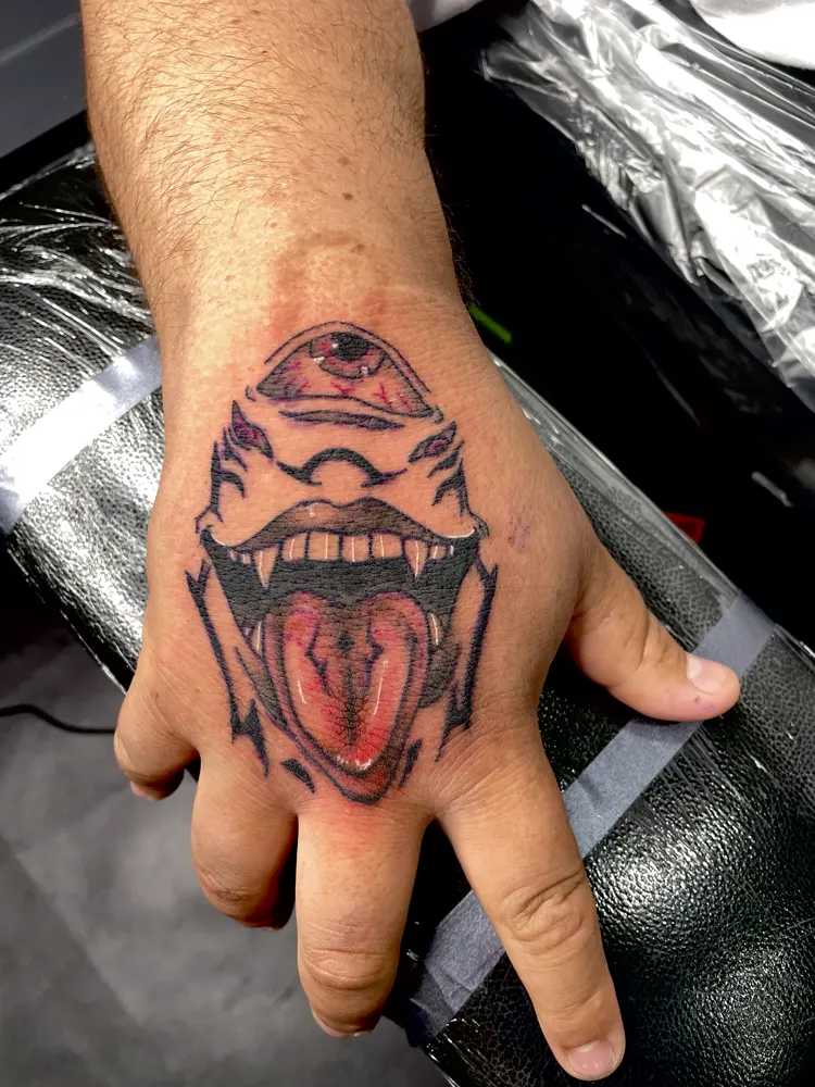 One-eyed monster tattoo with fangs and a long red tongue on the back of the hand