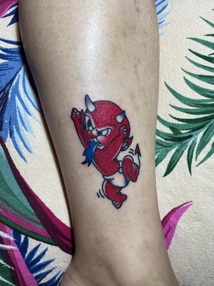 Red baby devil tattoo with horns and a pointed tail on the lower leg