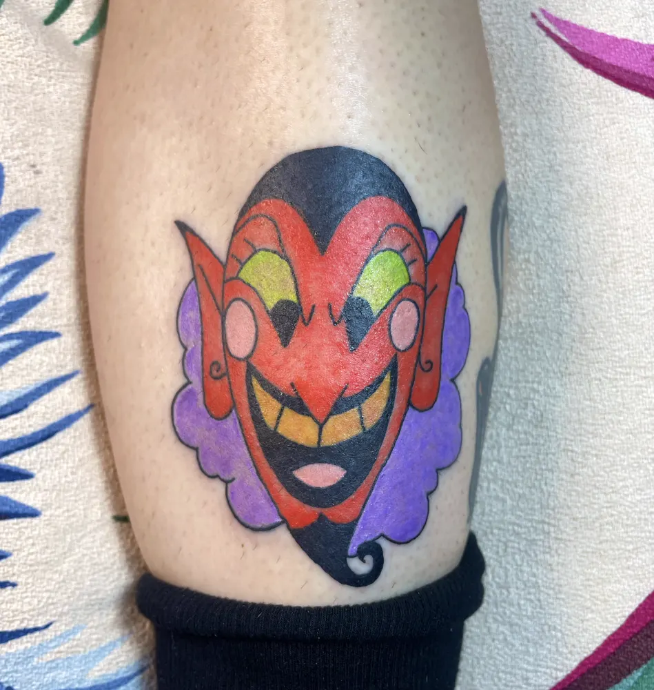 Grinning red devil head tattoo with yellow eyes on a purple cloud on the calf