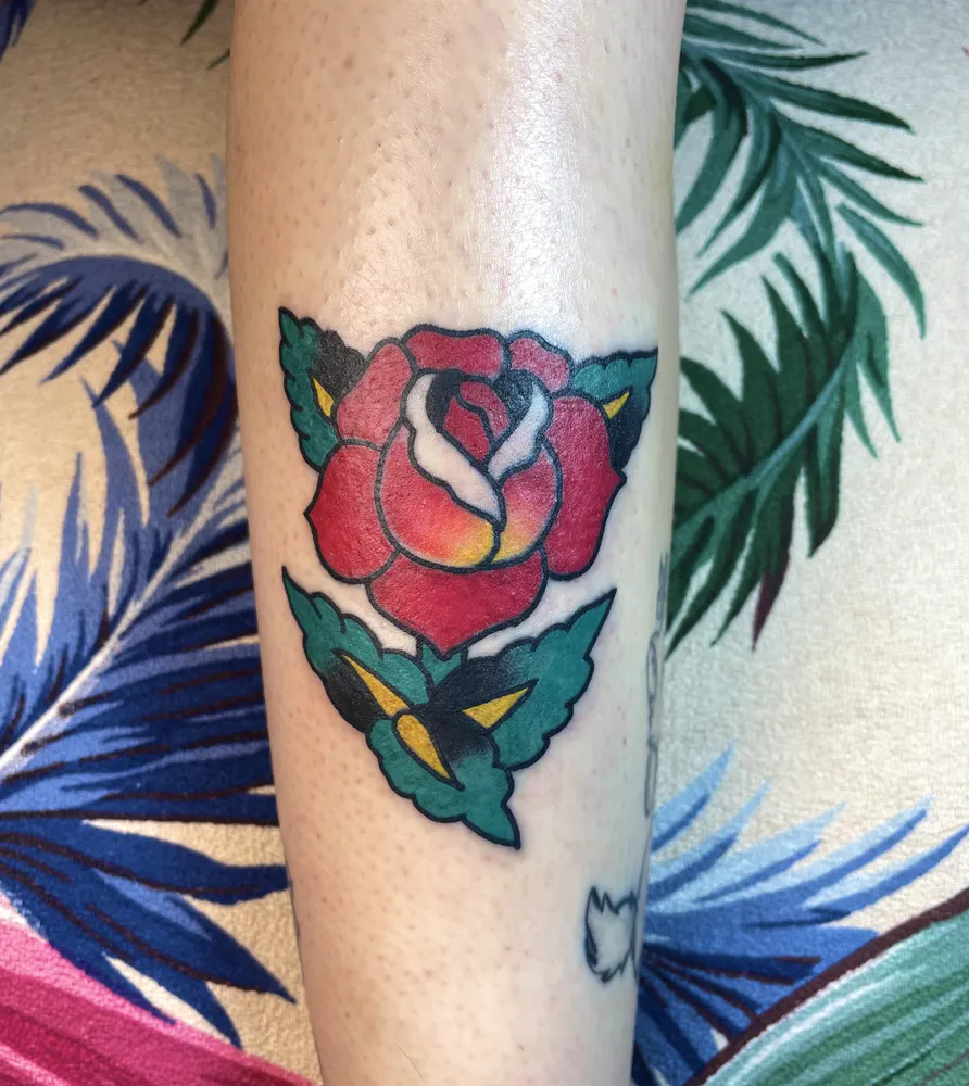Red rose tattoo with green leaves in American Traditional style on the shin
